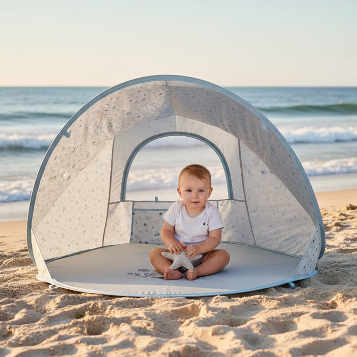 Baby sitzt im Schatten in einer blauen Strandmuschel am Strand mit Meer im Hintergrund – UV Schutz Pop Up Strandzelt für Babys und Kinder