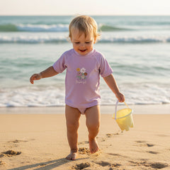 Baby läuft am Strand und trägt ein mauvefarbenes Bade-T-Shirt mit Blumenmotiv – sommerliche Baby-Bademode am Meer